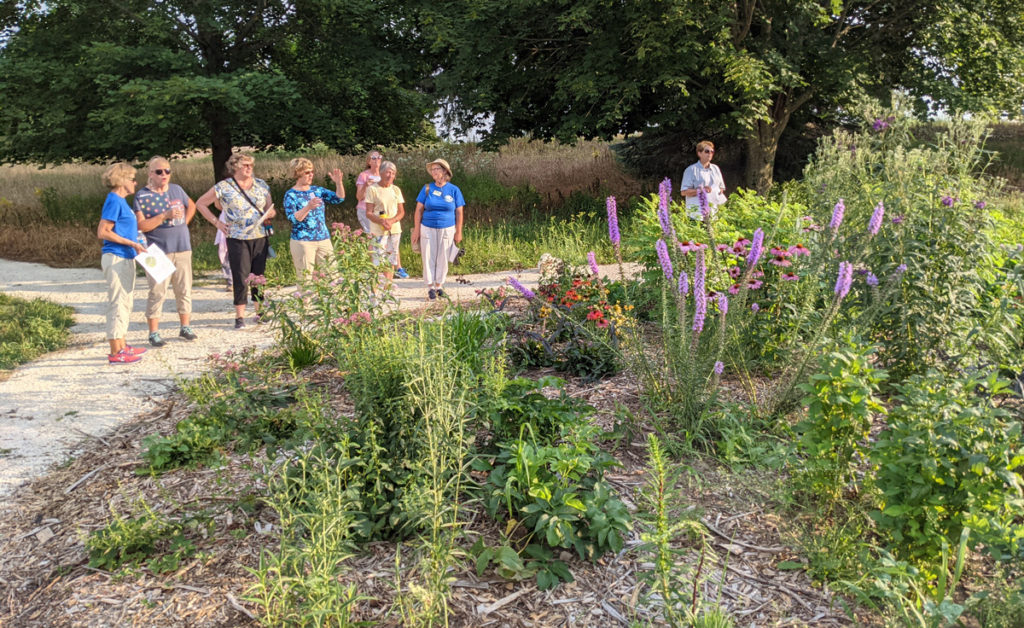A group of 8 people on a trail in front of a large perennial garden. There are trees in the background.