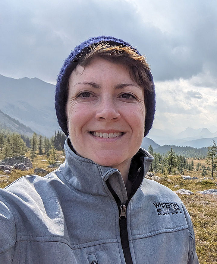 A middle aged woman wearing a knit cap, smiling in a selfie with mountains in the background
