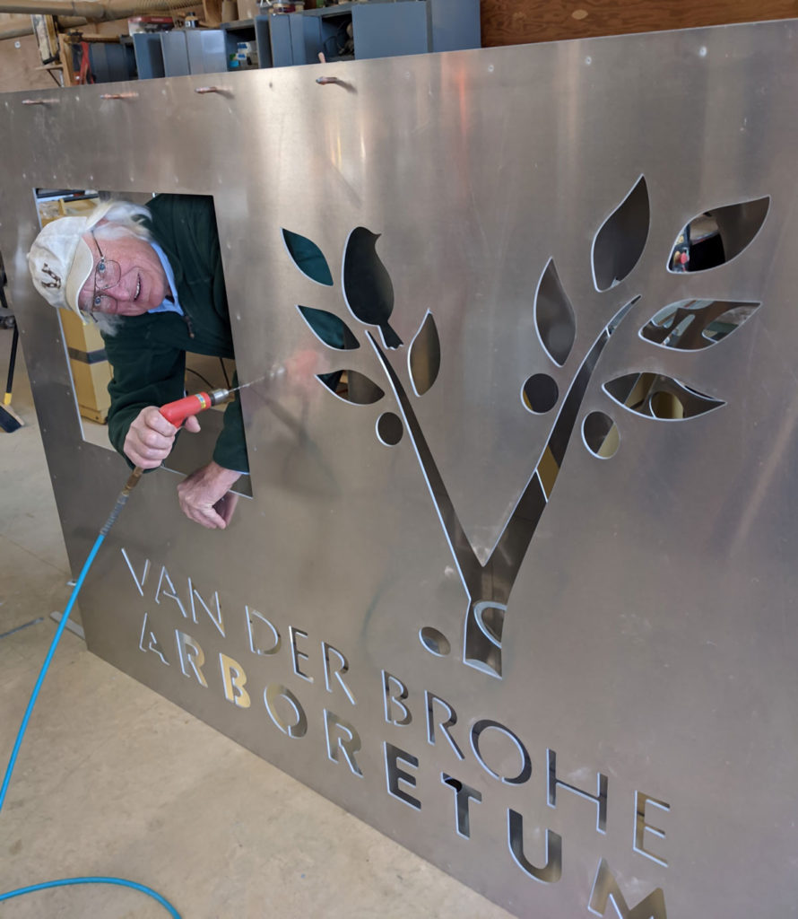 A man with white hair and a cap drills on a large aluminum sign while peeking through a cutout in the sign. The sign also has a tree and bird cutout, as well as the worlds VAN DER BROWH ARBORETUM.